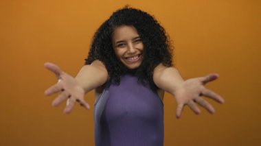 Canlı turuncu bir arka planda kollarını açmış, içten bir şekilde gülümseyen bir kadın basit bir ortamda dostluk ve neşe yayıyor..
