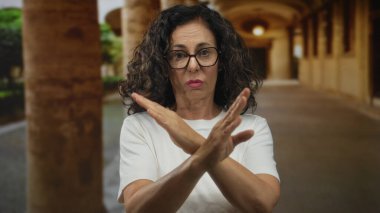 Orta yaşlı kıvırcık saçlı ve gözlüklü İspanyol kadın, eski kasaba sokaklarında reddedilme jesti olarak kollarını çaprazlıyor..