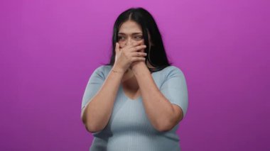 Kadın elleri pembe arka planda durarak ağzını kapatıyor, şaşkınlığını ifade ediyor, mavi gömlek giyiyor, duygularını ifade ediyor, genç, iri yarı bir kadına ait, canlı bir ortamda izole edilmiş..