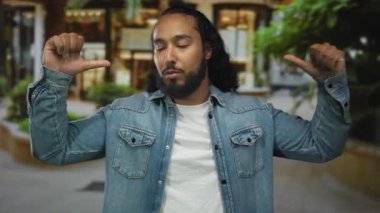 Kot ceketli bir adam, sakal ve kapalı gözlerle, sakallı ve gururlu bir şekilde, sakal ve bina yakınında, sokakta baş parmağıyla göğsüne işaret ediyor..
