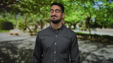 Gözlüklü genç İspanyol adam, güneşli bir parkta güvenle gülümsüyor yemyeşil ağaçları ve arka planda sakin bir atmosferi var.