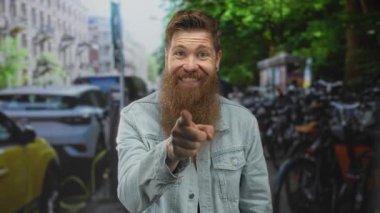 Uzun sakallı, kot ceket giyen bir adam park halindeki bisikletlerle dolu kalabalık bir şehirde parmaklarını dudaklarına bastırıyor..