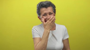 Gri saçlı yaşlı kadın neşeyle gülüyor ve ağzını kapıyor, canlı sarı bir arka plan karşısında duruyor, beyaz bir gömlek giyiyor, mutluluk ve pozitifliği ifade ediyor..