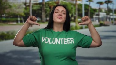 Yeşil gönüllü gömlekli genç bir kadın arka planda palmiye ağaçları olan güneşli bir caddede kendini göstererek gönüllü ruhunu açık havada sergiliyor..