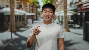 Beyaz gömlekli genç adam, güneşli bir şehir sokağında palmiye ağaçlarıyla eğlenceli bir jest yapıyor, açık hava kentsel manzarasını çekiyor..