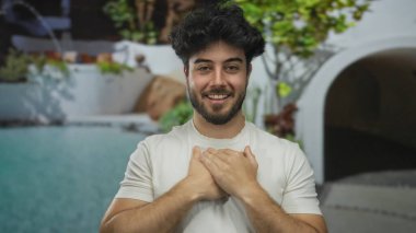 Sakallı genç bir adam otel ortamında havuz kenarında gülümsüyor, yemyeşil ve modern mimarinin ortasında rahat bir tatil havası taşıyor..