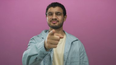 Stüdyoda pembe duvarlı, gözlüklü ve kot gömlekli bir adam kamerayı işaret ediyor..