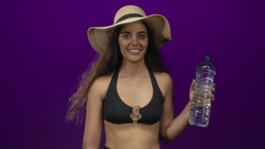 Genç İspanyol kadın elinde temiz bir su şişesi ve mor bir stüdyoda gülümsüyor..