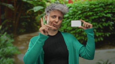 Kadın, orman ortamında yeşil yapraklara dayanan boş plastik kartı işaret ediyor; şüphecilik.