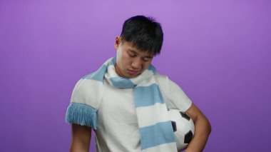Mor arka planda izole edilmiş futbol topunu tutan mavi eşarplı beyaz tişörtlü genç Çinli adam tefekkür ediyor..