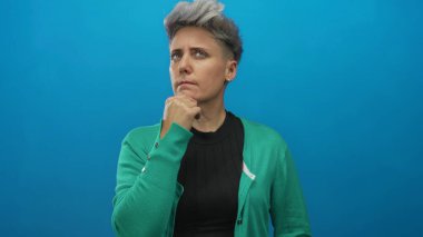 Mavi stüdyo duvarının önünde kısa saçlı ve eli çenesinde dalgın bir ifade olan bir kadın..