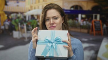Kadının elinde mavi kurdeleli, kapalı gözlü ve restoran terasında büzüşmüş dudakları olan bir hediye kutusu var..