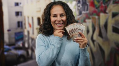 Orta yaşlı bir kadın Meksika pezosu tutarken gülümsüyor. Arkasında renkli graffiti olan canlı bir şehir caddesinde para ve şehir hayatı sergiliyor..