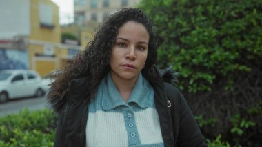 Parkta duran bir kadın, ciddi bir ifadeyle, kentsel bir ortamda kışlık ceket giyiyor, arka planda ağaçlar ve binalar görünüyor..