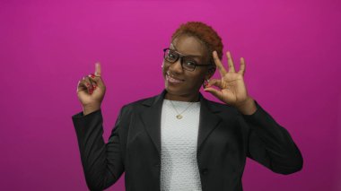 Gözlük ve halka küpe takan bir kadın, parlak pembe bir stüdyo geçmişiyle el işareti yapıyor. Onaylandı..