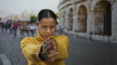 Sarı kazaklı kadın Roma Kolezyumu 'nda parmak tabancası hareketi yapıyor. Arka planda antik mimari ve kaldırım taşı caddeleri var..