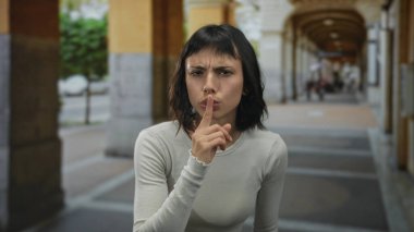 Genç İspanyol kadın sessiz kalmak için parmaklarını dudaklarına uzatarak tarihi bina kemerlerinin altında güneş ışığıyla aydınlanan sokak sütununda duruyor..
