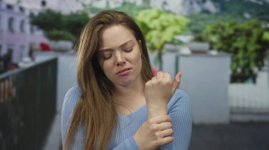 Gün ışığında bulanık bir sokak cephesine karşı tek eliyle bileğini tutan bir kadın..