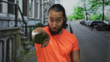 Turuncu gömlekli adam, park halindeki arabaları ve tuğla binaları olan şehir caddesindeki kamerayı işaret ediyor..