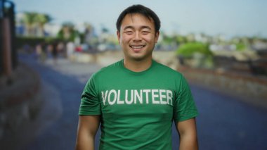 Yeşil gönüllü tişörtü giyen gülümseyen adam şehir sokaklarının arka planında duruyor. Şehir ortamını ve toplum ruhunu gözler önüne seriyor..