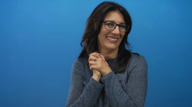 Gözlüklü orta yaşlı kadın mavi stüdyoda elleri sımsıkı bağlı gülümsüyor; minnettarlık içtenliği.