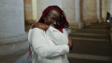 Örgülü saçlarıyla gülümseyen kadın, arka planda mimari sütunlar bulunan kilise ortamında kendini kucaklıyor..