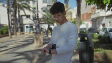 Palmiye ağaçlarının yanındaki güneşli yeşil bir parkta akıllı telefon kullanan ve bileklerine tıklayan bir adam; farkındalık yansıması iç gözlem odağı.