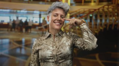 Gülümseyen kısa saçlı kadın lüks otel lobi resepsiyonunda yanak yanağa parmakla işaret ediyor; güven güçlendirme hırsı.