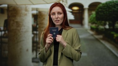 Kızıl saçlı kadın elinde Alman pasaportuyla şehir caddesinde düşünceli ve düşünceli bir şekilde büyük bir tepe örtüsünün altında duruyor..