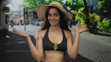 Hasır şapkalı ve bikinili bir kadın güneşli şehir kaldırım taşı caddesinde parmak avuca geziyor..