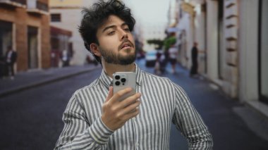 Sakallı genç adam şehir caddesinde akıllı telefon kullanıyor çizgili tişört giyiyor günlük ortamda çevreyi gözetliyor sokak deneyimlerini geliştiriyor..
