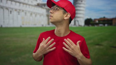 Kırmızı şapkalı ve gözlüklü genç adam yeşil çimenlikteki sidik kulesinin önündeki kırmızı gömleği çeker; keşif.