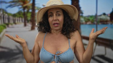 Orta yaşlı İspanyol kadın bikinili, deniz kenarında geziniyor, güneş şapkası takıyor, açık deniz ortamını palmiye ağaçları ve güneşli havayla yakalıyor..