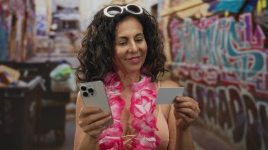 Bikini giyen ve Ley takan bir kadın akıllı telefon ve kredi kartını dışarıda duvar yazısının karşısında tutuyor..