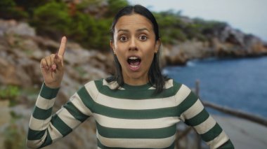 Genç İspanyol kadın, güneşli bir açık havada, okyanus ve doğa tarafından çevrelenmiş neşeli bir ifadeyle manzaralı bir sahilde dikilirken fikir jesti yapıyor..