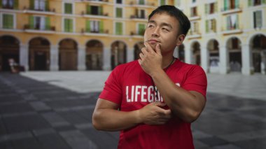 Kırmızı tişörtlü genç Çinli bir cankurtaran eski kasaba caddesinde kolları çapraz olan bir düdük kolyesi takıyor..