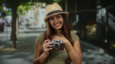Açık havada gülümseyen bir kadın elinde eski model bir kamerayla güneşli bir sokakta neşe saçan ve şehir ortamında seyahat anılarını yakalayan bir şapka takıyor..