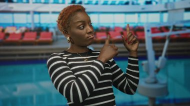 Kırmızı tırnaklı, çizgili siyah kazaklı kadın bina içindeki nesneyi işaret ediyor; şüphecilik.