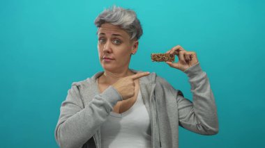 Kısa saçlı, yüzü buruşmuş ve parmakları turkuaz stüdyoda atıştırmalık olduğunu gösteren granola çubuğu tutan bir kadın..