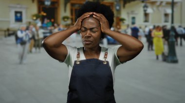 Önlüklü bir kadın ellerini başının üstüne koyup, stresi ifade ediyor. Etrafı insanlarla çevrili ve kentsel hayattaki zorluklarla dolu..