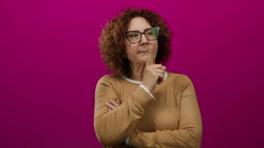 Gözlüklü kadın canlı pembe bir arka plana karşı şşşş hareketi yapıyor, kahverengi bir kazak giyiyor ve zarafet ve gizem saçıyor..