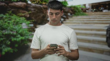 Beyaz gömlekli adam iki elinde akıllı telefon tutuyor ve yeşil yapraklı merdivenlere yan bakıyor; merak araştırma bağlantısı.