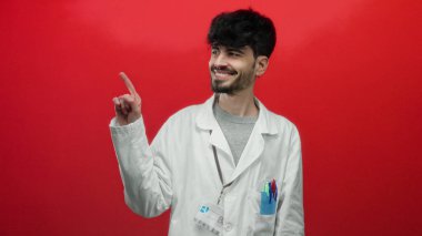 Beyaz önlüklü, parlak kırmızı duvara karşı rozeti olan genç bir adam, muhtemelen profesyonel ya da tıbbi bağlamda, kendine güvenerek işaret ediyor..