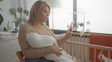 Stüdyoda akıllı telefon ekranını kaydırırken kadın örgü battaniyeye sarılmış bebeği tutuyor; annelik huzuru.