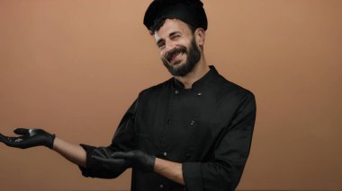 Siyah şef üniformalı İspanyol adam, turuncu bir arka plana karşı el uzatıyor. Mutfak uzmanlığını ve konukseverliğini hoş bir jestle yakalıyor..