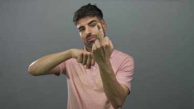 Pembe polo tişörtlü genç İspanyol adam gri stüdyoda kameraya bakan orta parmağını gösteriyor..