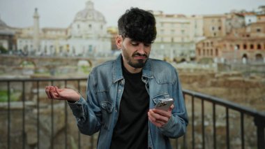 Kot ceketli genç adam Roma harabelerinde dikiliyor telefon ekranına bakıyor ve tarihi şehrin arka planına kafa karışıklığını dile getiriyor..