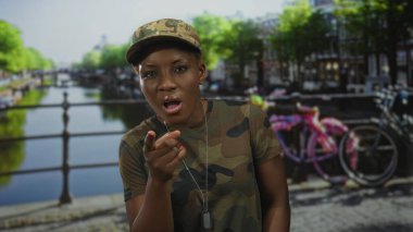 Kamuflaj üniformalı kadın asker köpek etiketi kolyesiyle ağzını açar ve parmağıyla sokağı işaret eder; acil uyarı.