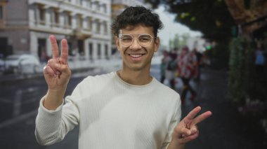 Beyaz süveter giyen ve gözlüklü bir adam iki eliyle sokak köşesinde barış işareti yapıyor. İyimserlik..