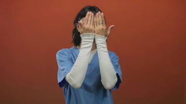 Genç kadın hemşire gülümserken yüzünü kırmızı bir stüdyo duvarına karşı açık avuçlarla kapatır..